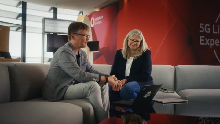 A man and woman seated on a couch in a professional setting. They smile while watching something on a laptop. Behind them is a red wall with a 